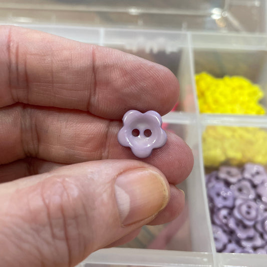 Hand holding a small purple flower-shaped button with a clear container of buttons in the background.