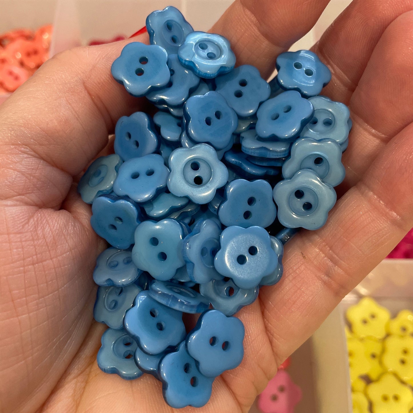 Hand holding blue flower-shaped buttons with a container of more buttons in the background.