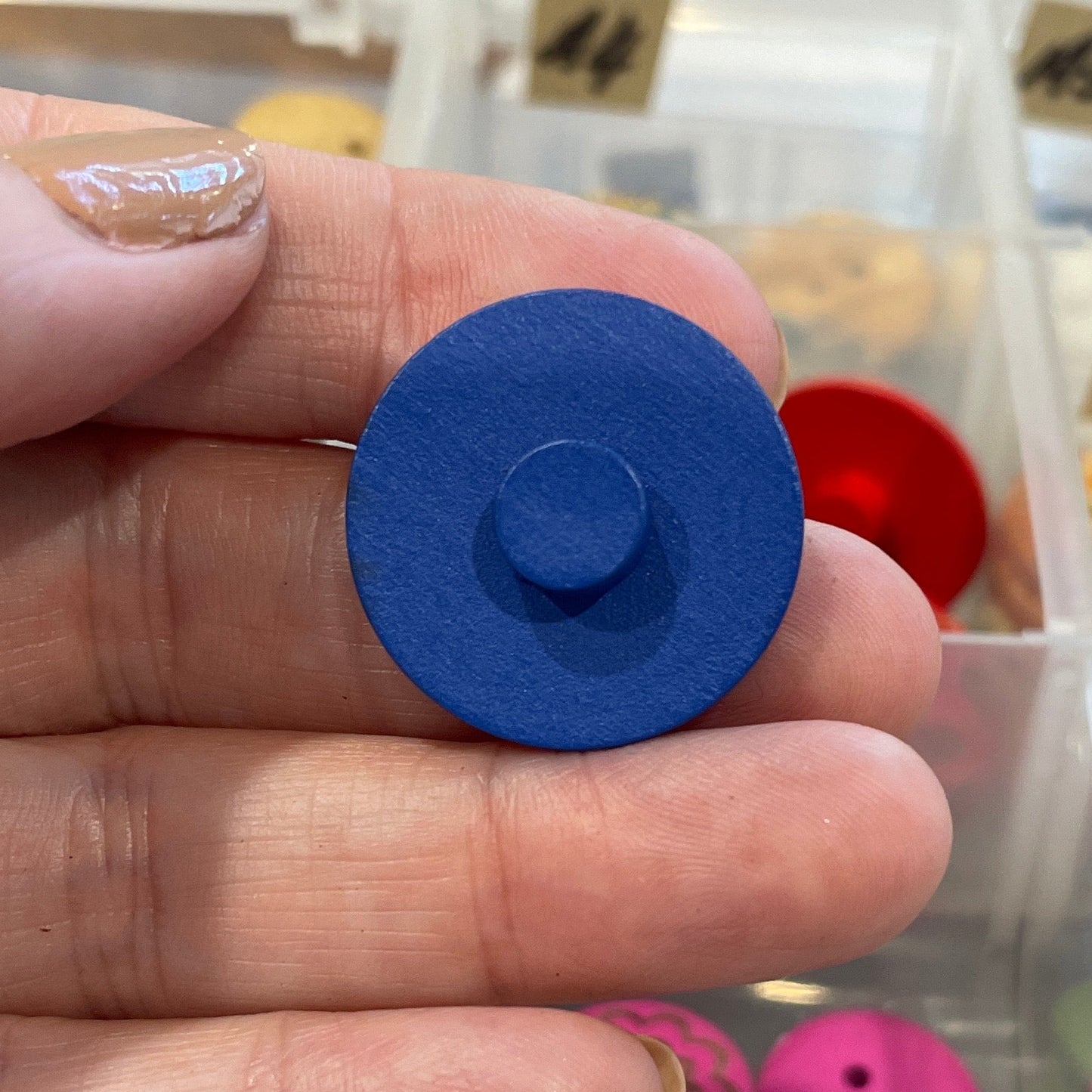 Hand holding a blue button with a collection of buttons in a clear container in the background