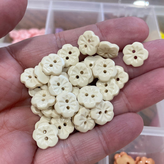 Hand holding a small pile of white flower-shaped buttons with a blurred container of buttons in the background.