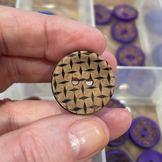 Hand holding a wooden button with a pattern, with a container of buttons in the background.