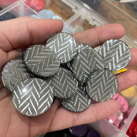 Hand holding silver textured buttons with a container of colorful buttons in the background.