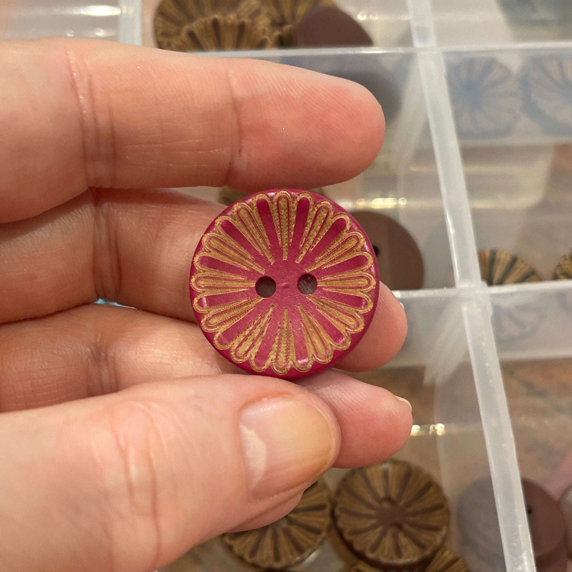 Hand holding a pink floral button with a clear container of assorted buttons in the background