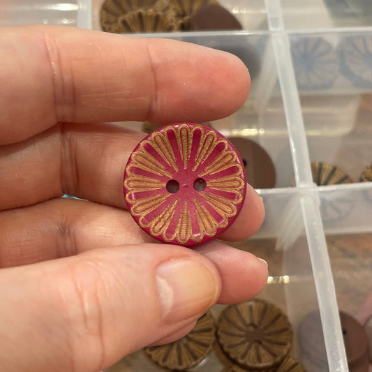 Hand holding a pink floral button with a clear container of assorted buttons in the background