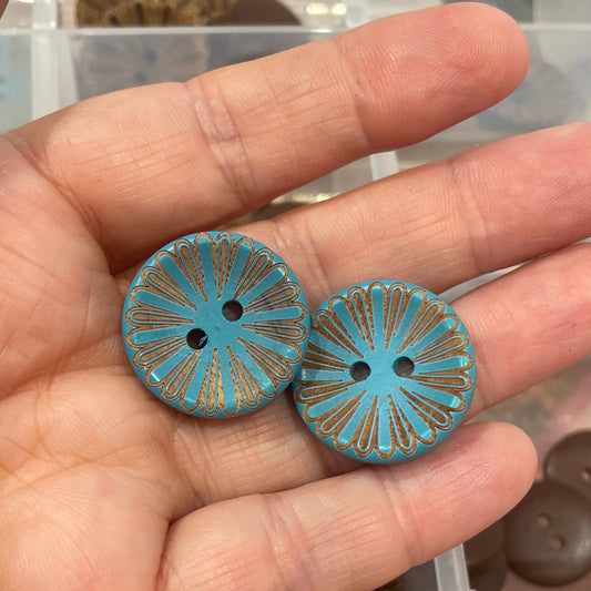Hand holding two blue decorative buttons with a container of assorted buttons in the background.