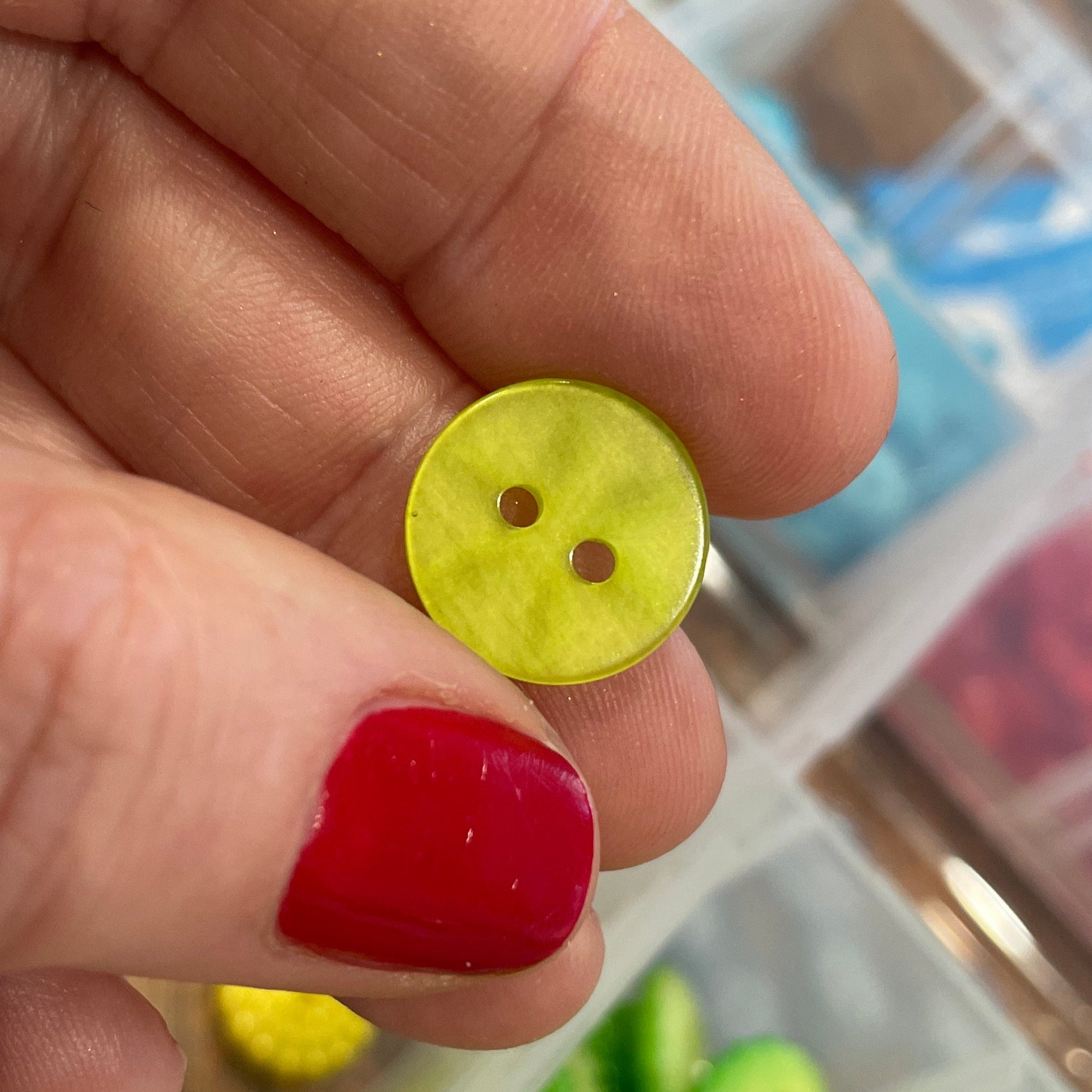Hand holding a small yellow button with two holes, with a blurred background of colorful items in containers.