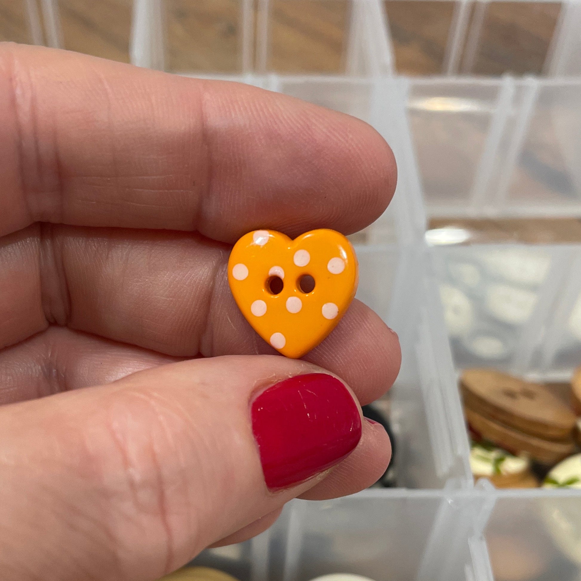 Hand holding a yellow button with a heart design in front of a container of buttons.