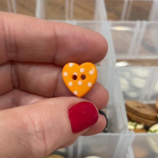 Hand holding a yellow button with a heart design in front of a container of buttons.