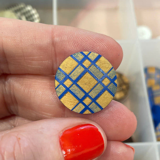Hand holding a blue and gold checkered button with a container of buttons in the background