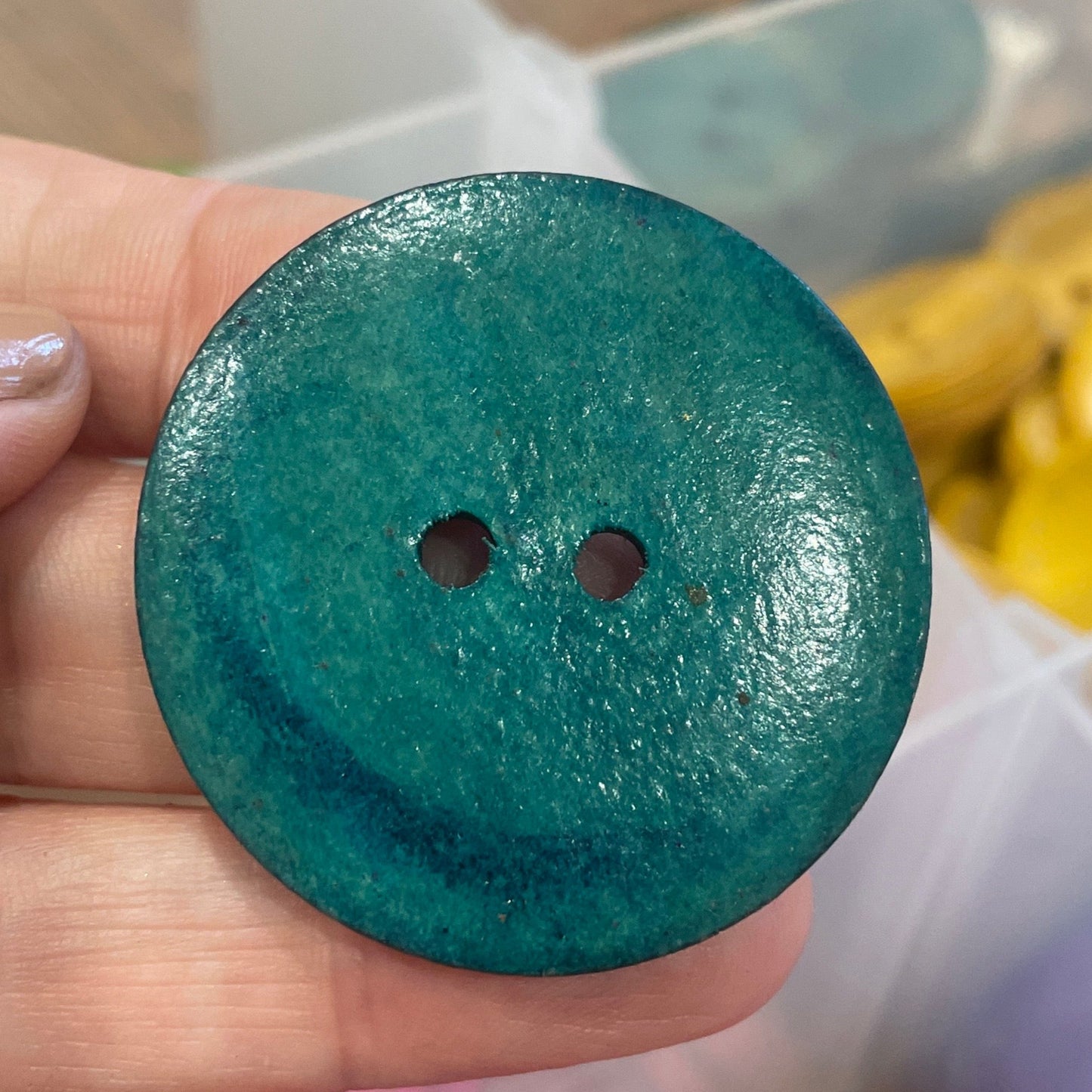 Hand holding a teal button with a container of colorful buttons in the background
