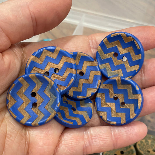 Hand holding blue and gold zigzag patterned buttons with a container of similar buttons in the background.