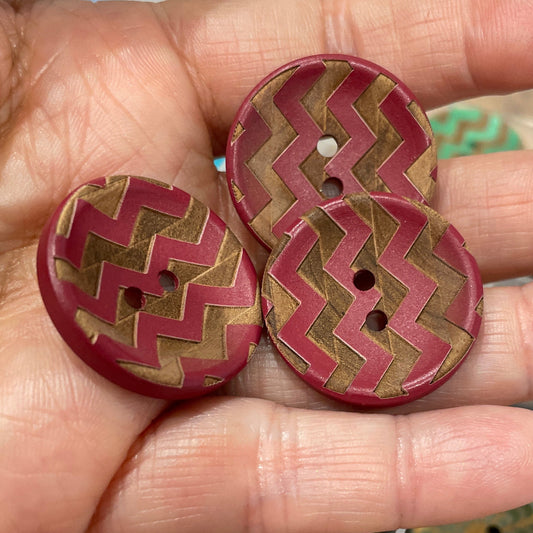Hand holding three decorative buttons with a zigzag pattern, with a container of more buttons in the background.