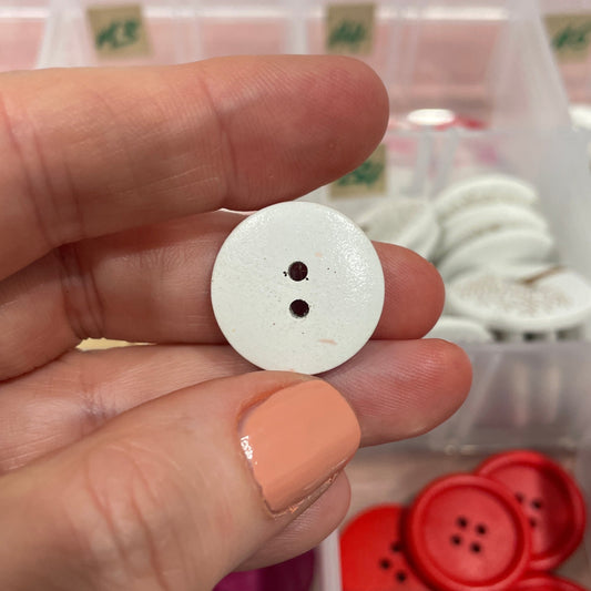 Hand holding a white button with a container of various buttons in the background