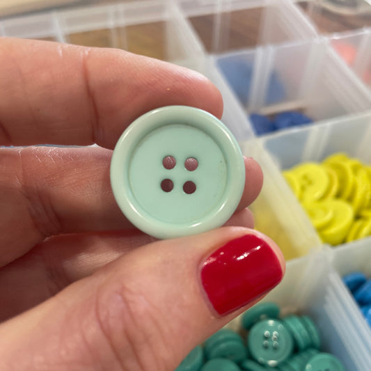 Hand holding a light green button with a container of colorful buttons in the background