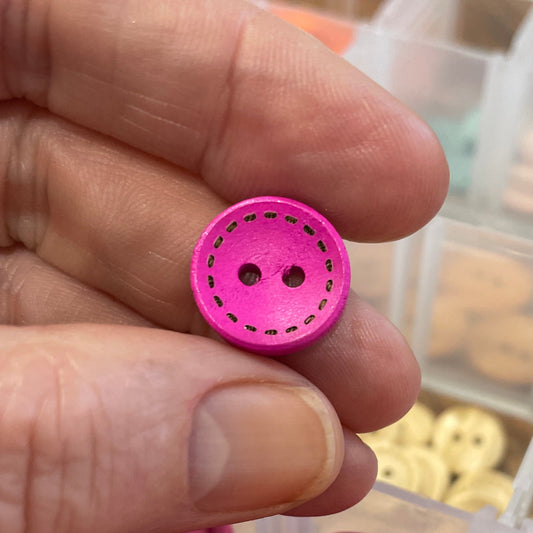 Hand holding a pink button with a container of assorted buttons in the background
