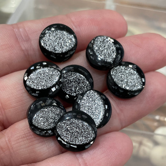 Hand holding black and silver glittery buttons with a container of red buttons in the background.