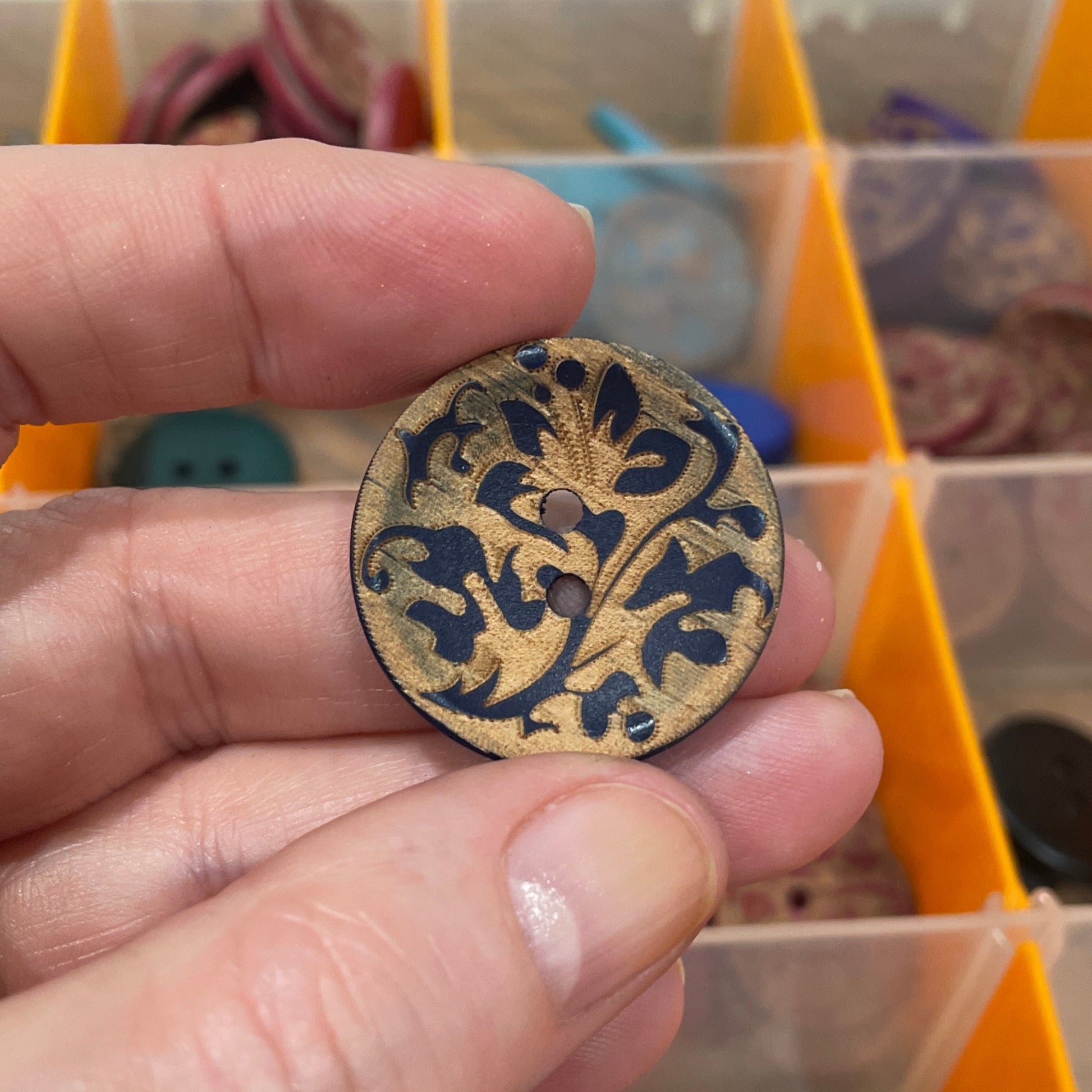 Hand holding a decorative button with a detailed design in front of a button organizer.