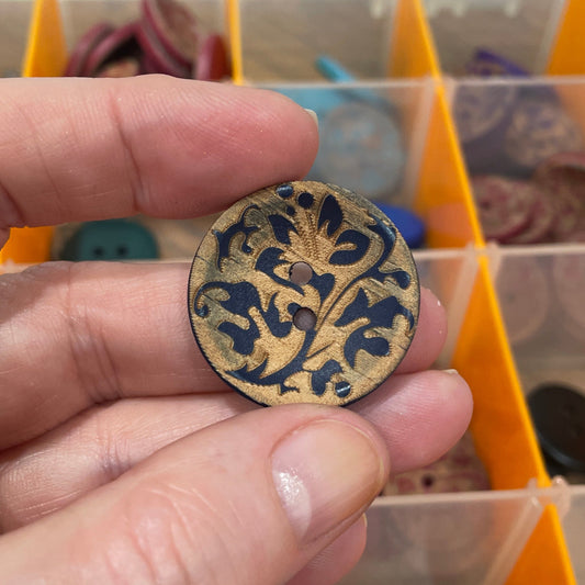 Hand holding a decorative button with a detailed design in front of a button organizer.