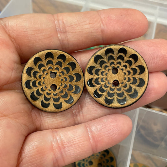 Hand holding two decorative wooden buttons with intricate designs, with a container of similar buttons in the background.