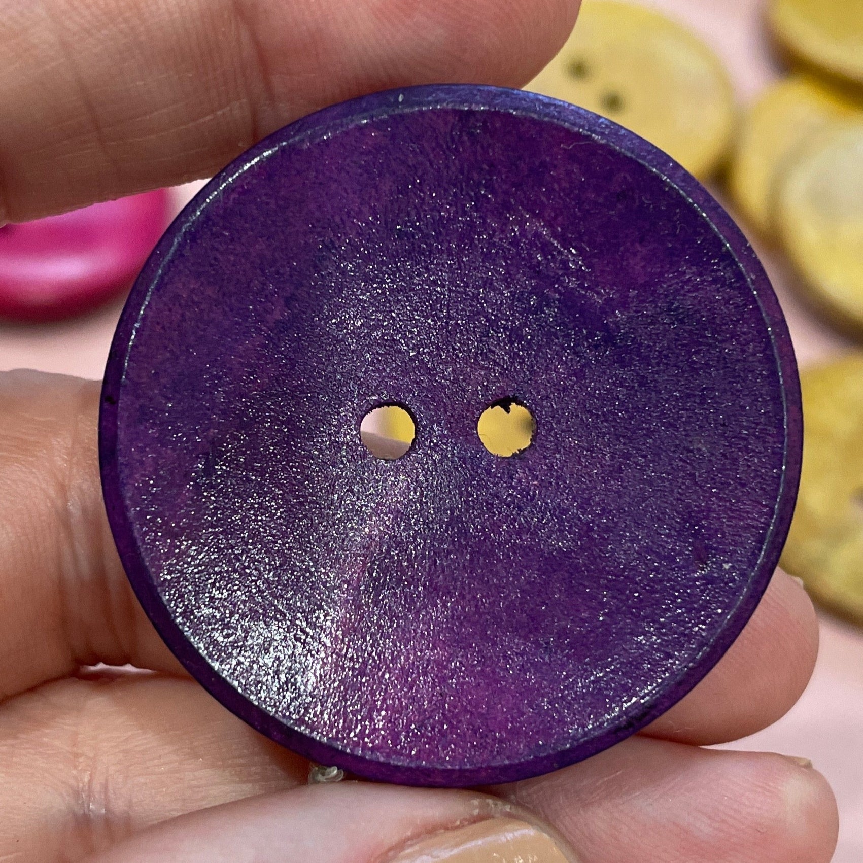 Hand holding a purple button with two small holes, surrounded by other buttons on a pink surface.