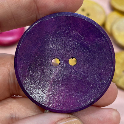 Hand holding a purple button with two small holes, surrounded by other buttons on a pink surface.