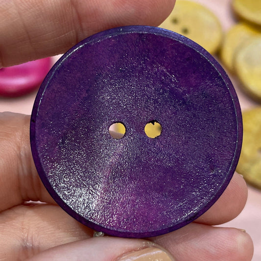 Hand holding a purple button with two small holes, surrounded by other buttons on a pink surface.