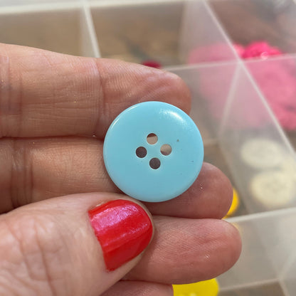 Light blue button held by a hand with red nail polish, against a background of colorful buttons in a clear container.