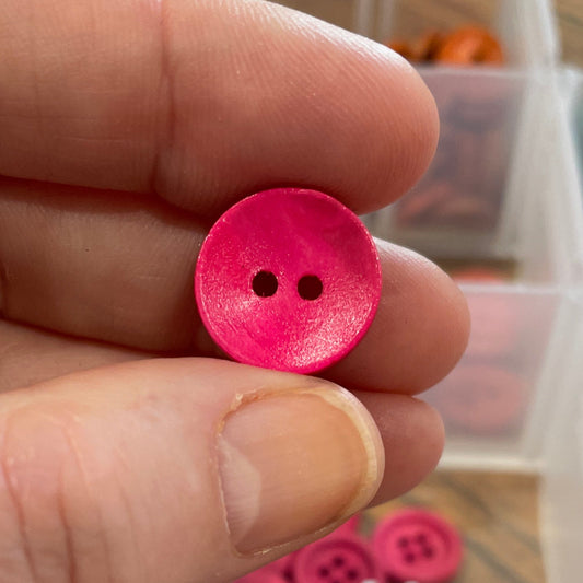 Hand holding a pink button with a container of assorted buttons in the background