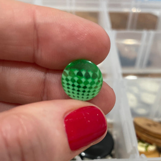 Green button held by a hand with red nail polish, against a background of assorted buttons in a container.