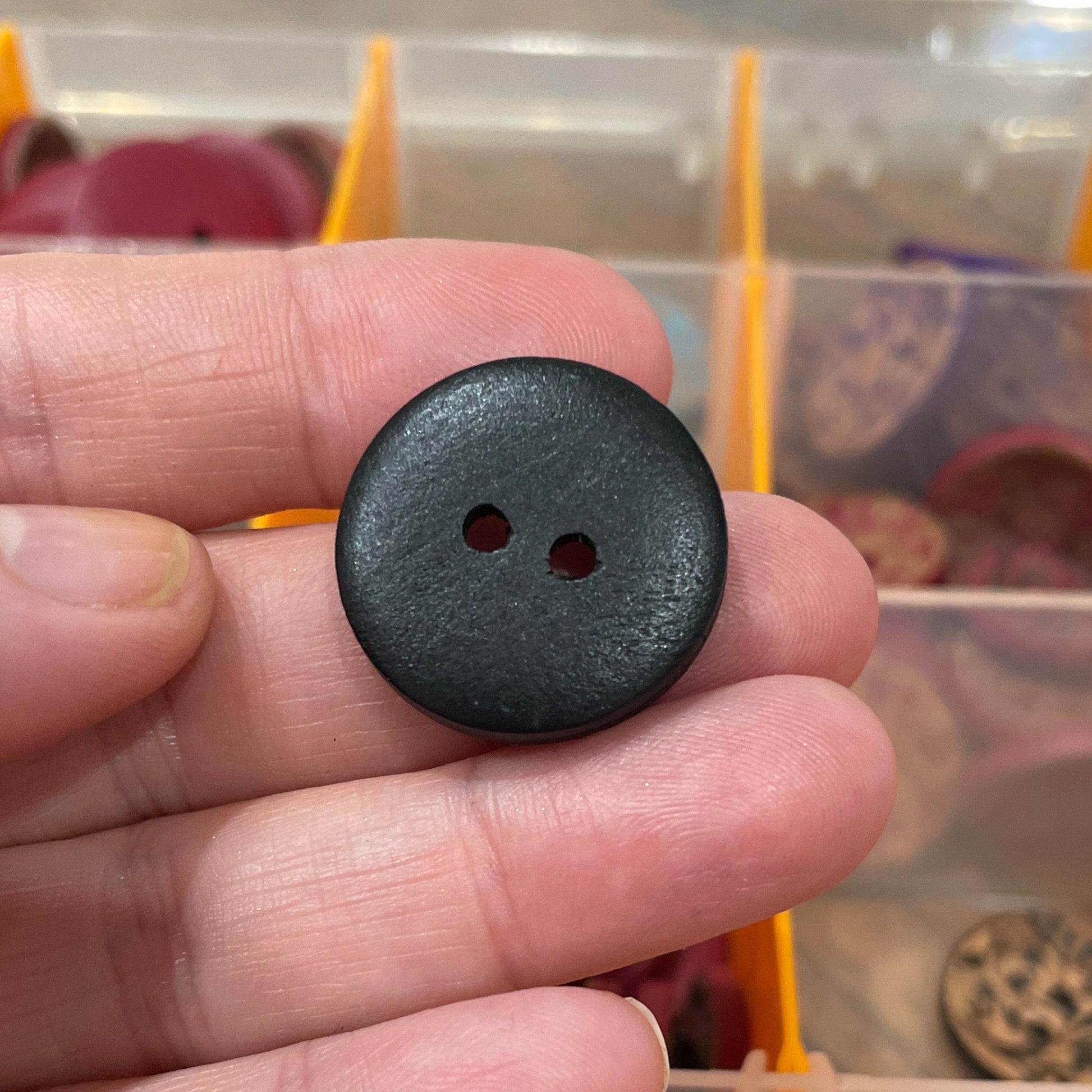 Black button held by a hand with a container of buttons in the background