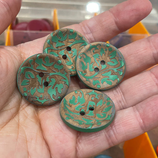 Hand holding four green buttons with floral patterns against a blurred background of a storage container.