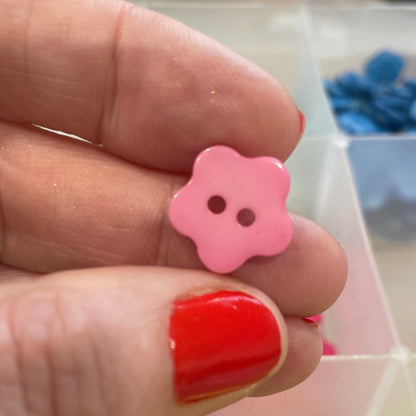 Pink flower-shaped button held by a hand with red nail polish, against a background of colorful buttons.