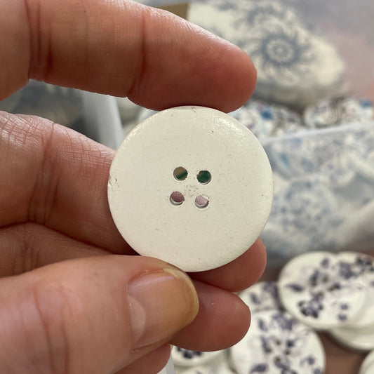 Hand holding a white button with three holes, with a container of similar buttons in the background.
