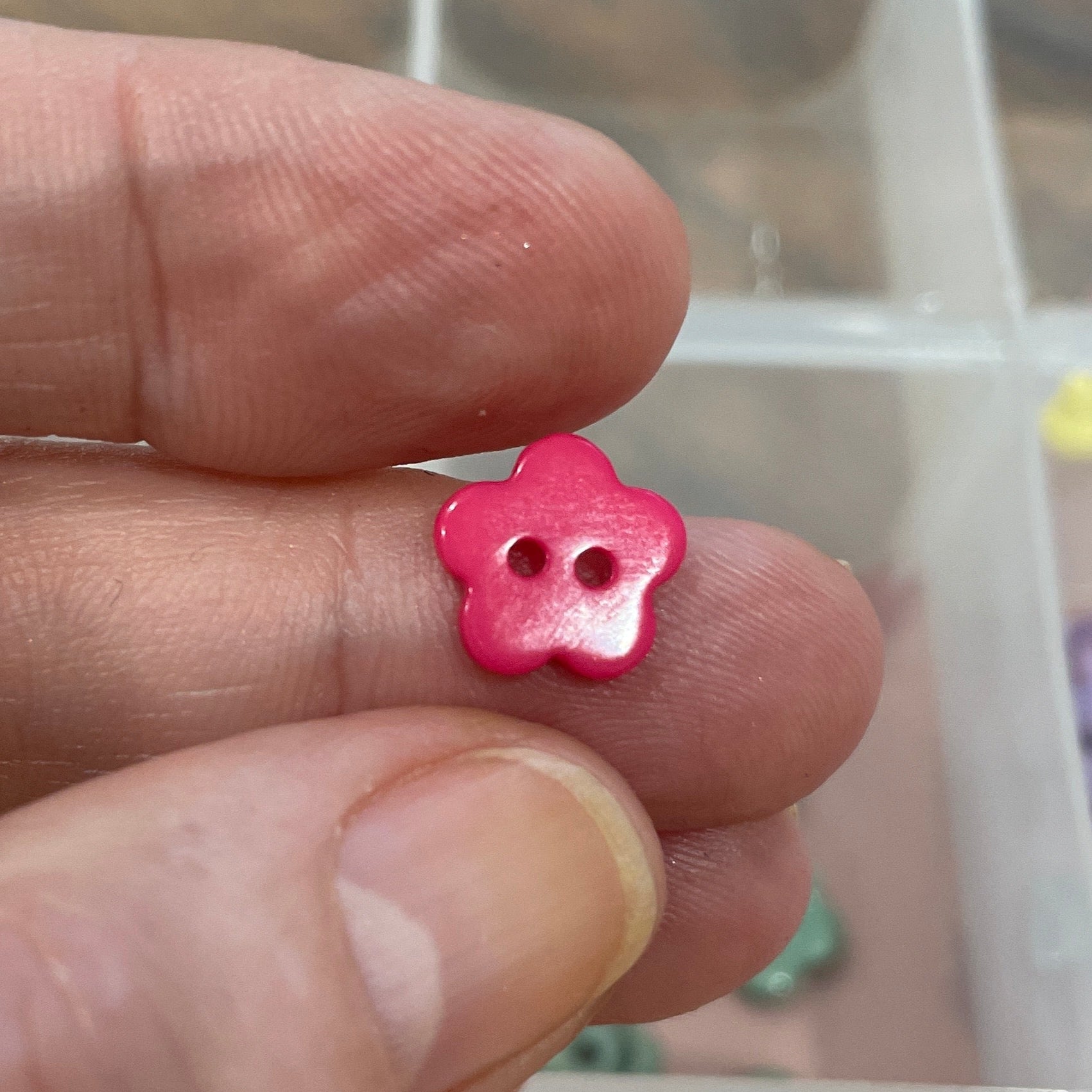 Pink flower-shaped bead held between fingers with a container of assorted beads in the background.