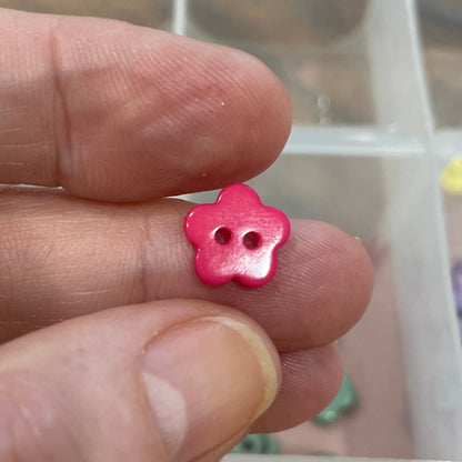 Pink flower-shaped bead held between fingers with a container of assorted beads in the background.