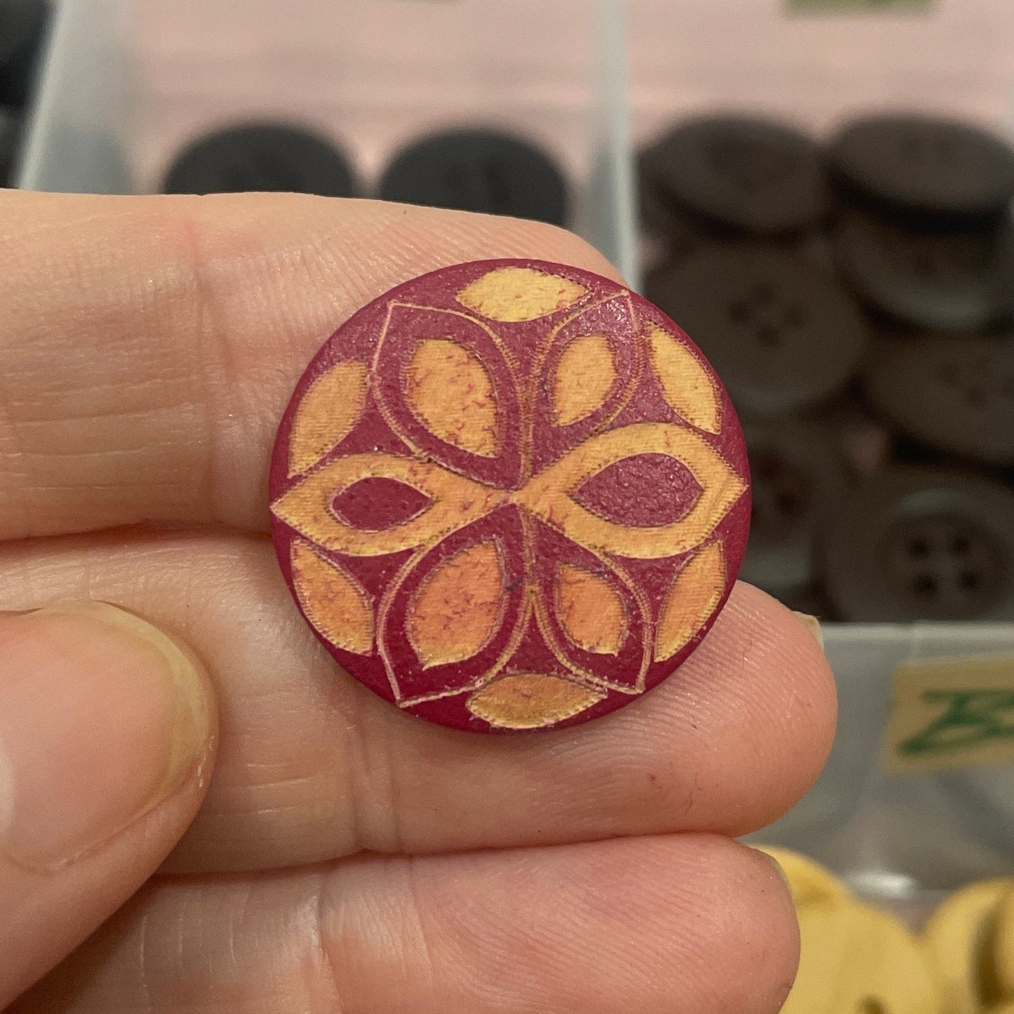 Hand holding a decorative button with a floral pattern in front of a container of assorted buttons.