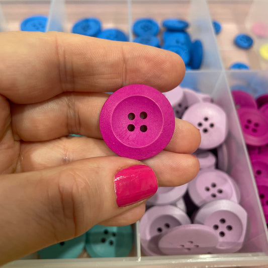 Hand holding a pink button with a container of assorted buttons in the background