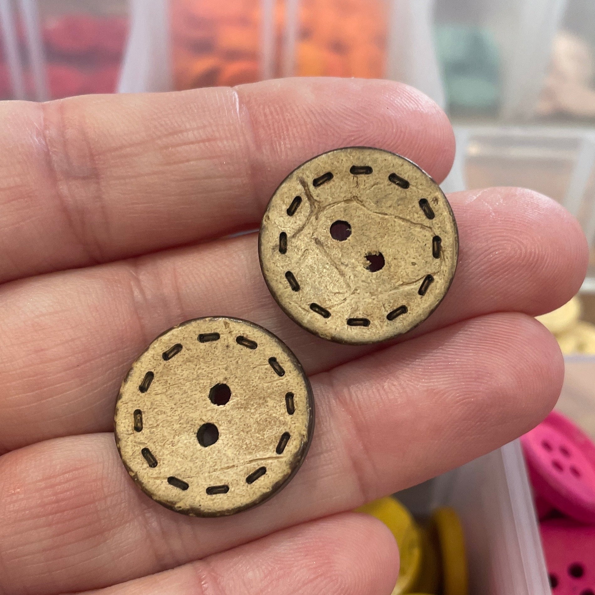 Two buttons held in a hand with a container of buttons in the background.