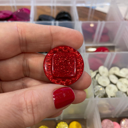 Hand holding a red glittery object with a container of colorful round objects in the background.