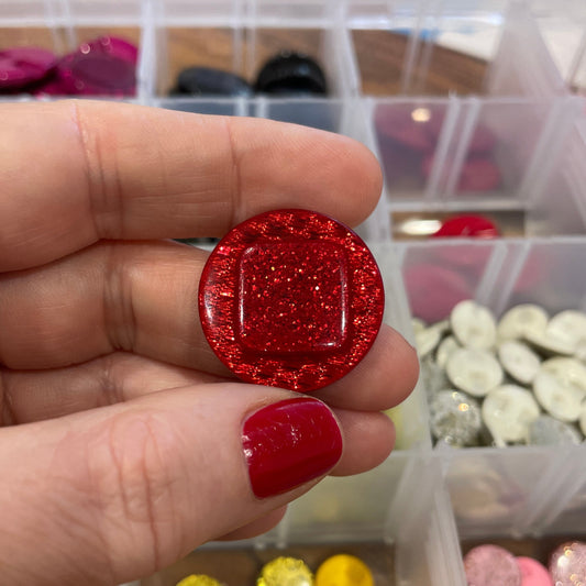 Hand holding a red glittery object with a container of colorful round objects in the background.