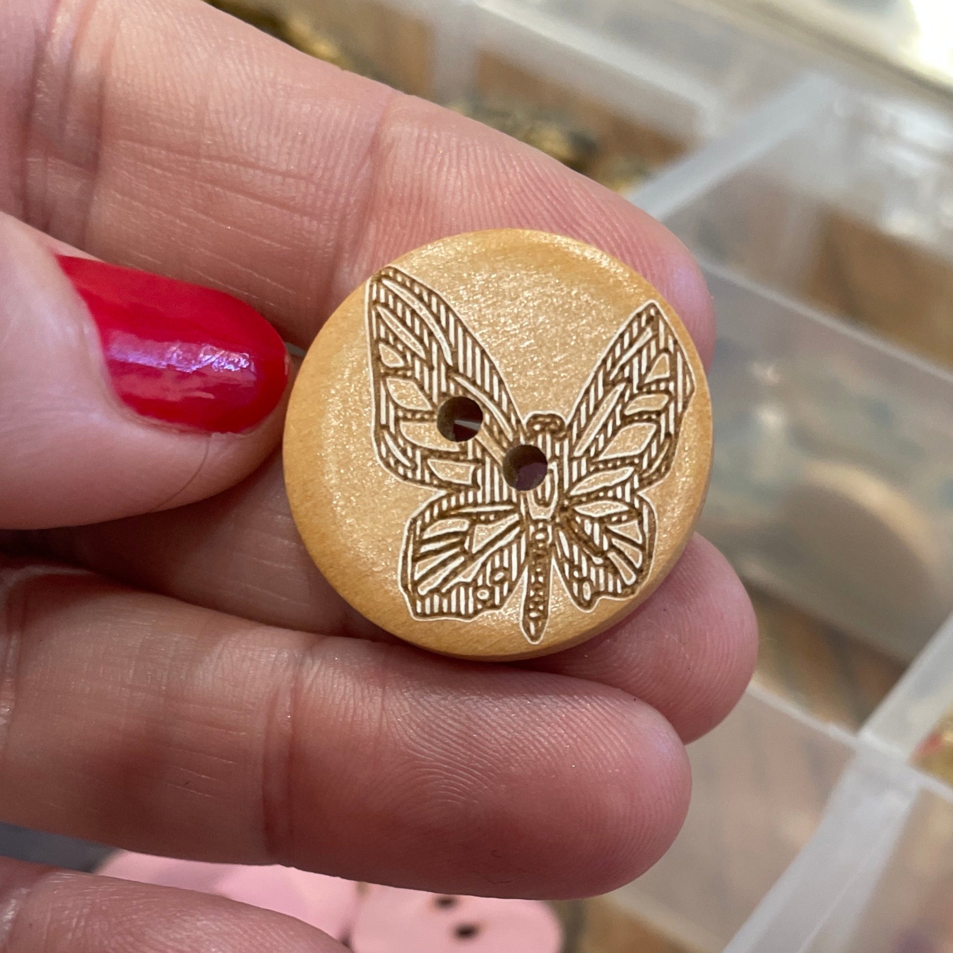 Wooden button with design held by a hand, with a container of colorful buttons in the background.