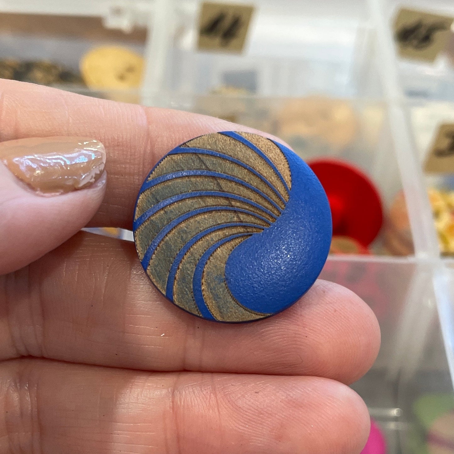 Hand holding a blue and gold decorative button with a background of various buttons in a clear container.