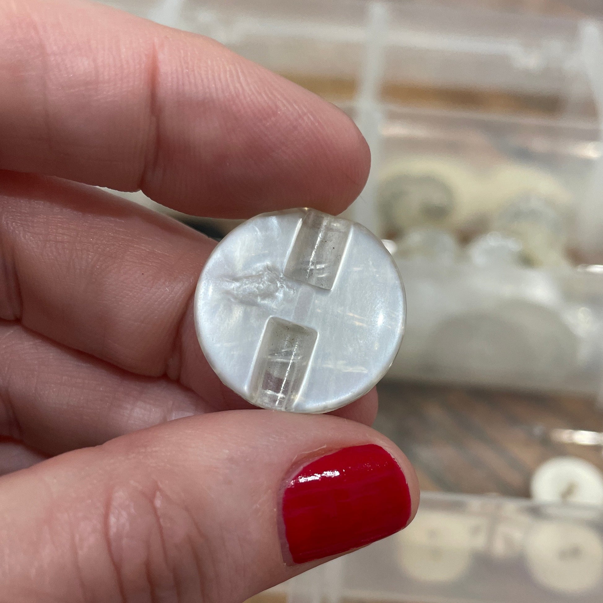 Hand holding a small round object with a clear plastic cover, against a background of buttons in a container.