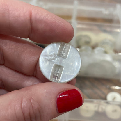 Hand holding a small round object with a clear plastic cover, against a background of buttons in a container.