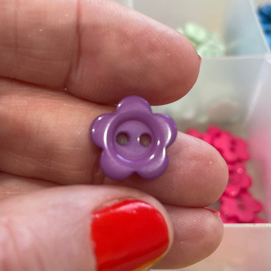 Hand holding a purple flower-shaped button with a container of colorful buttons in the background.
