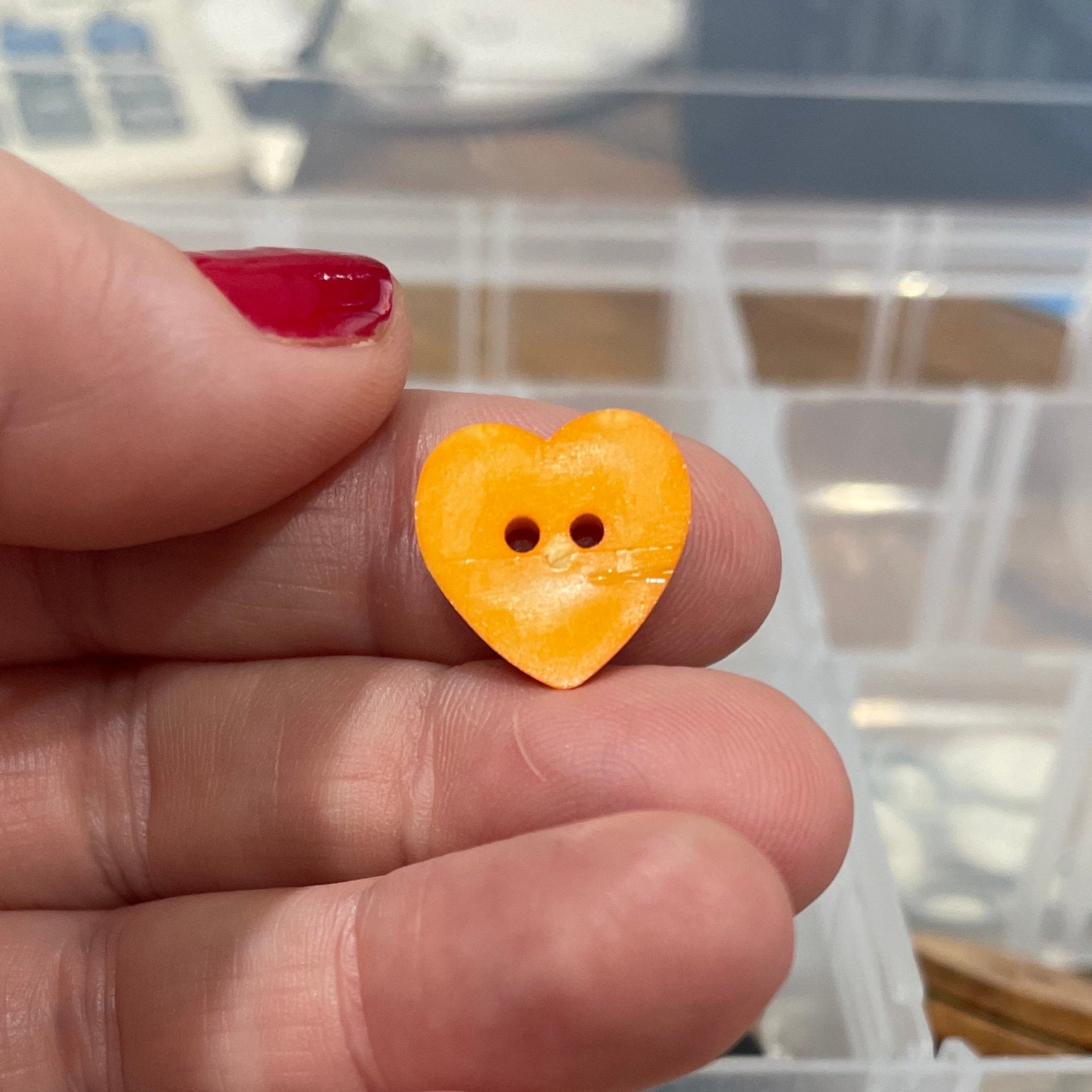 Hand holding a yellow heart-shaped button with a clear container of assorted buttons in the background.