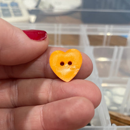 Hand holding a yellow heart-shaped button with a clear container of assorted buttons in the background.