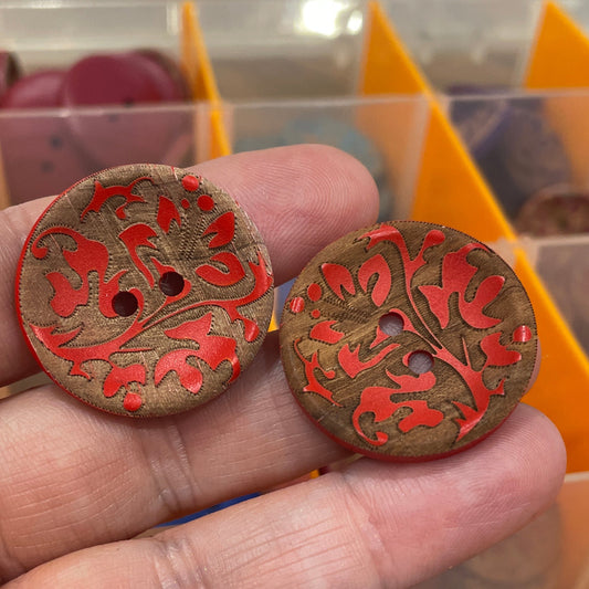 Two decorative buttons with red and gold patterns held by a hand, with a blurred background of shelves.