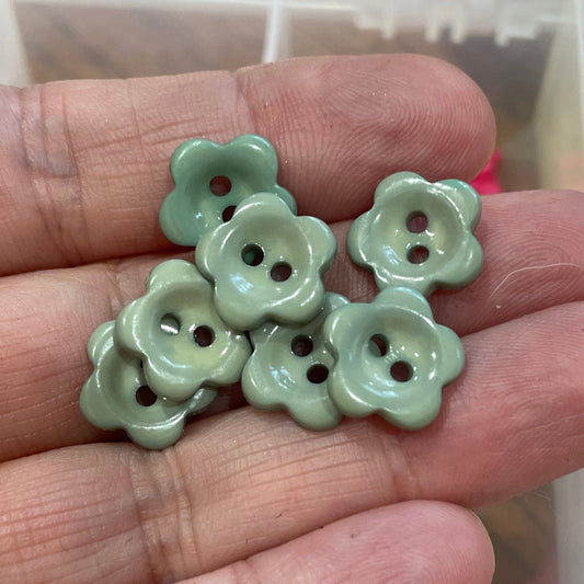 Hand holding green flower-shaped buttons with a container of assorted buttons in the background.