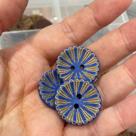 Hand holding two blue decorative buttons with a container of various buttons in the background.